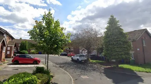 A number of semi detached and terraced brick properties on Dean Brook Close, with several leafy trees