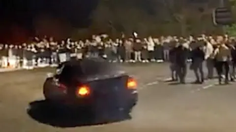 A car doing donuts around a group of people at a car meet