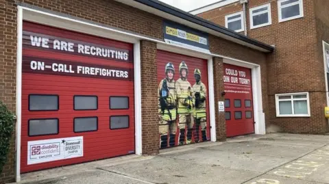 The exterior of Slade Park Fire Station with an empty forecourt. The garages are closed. A mural of three firefighters adorns one of the garage doors