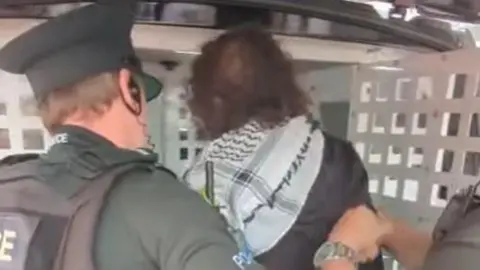 OTHER Screengrab of dark haired woman being loaded into the back of a police van. She is wearing a black and white scarf, probably a keffiyeh, and has a dark coat on. Two police officers, one more visible, are helping her into the van.