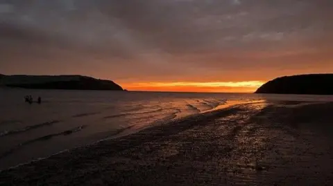 Shanti Nelson An orange and pink sky as the sun rises over a beach and the sea in the Black Isle - on the left hand side a few shadowy figures can be seen, heading out to sea