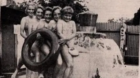 Shere Swimming Pool Club A black and white image of five children lined up smiling at the camera. They are stood next to what appears to be a water feature at a swimming pool. One is holding a large inflatable rubber ring
