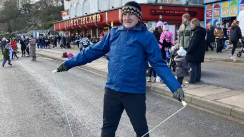 Freya Cox/BBC Malcolm Warnes stands with a skipping rope in the middle of a road. He wears a blue waterproof coat, a brown woolly hat and dark trousers. Behind him, an amusement arcade and groups of families and people can be seen. 