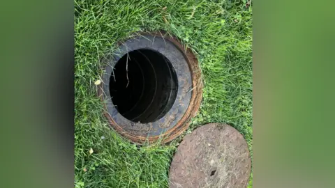 An open manhole with the cover next to it on the grass