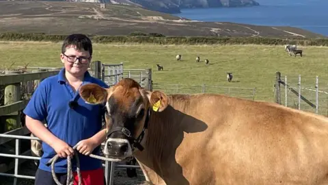 Charlie is wearing shorts, a blue T-shirt and a pair of glasses as he stands in a field in front of some metal fencing. He is holding a rope which leads to a bridle worn by a large brown Jersey cow next to him, Clare. There are sheep in the field behind them and beyond that the sea.