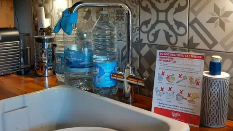 A tap with the paper warning not to use the tap water next to it. There is a blue glove tied around the end of the tap and a bottle of water on the right of the image.