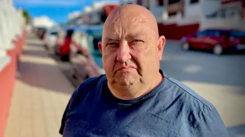 Billy George stands in the middle of the street looking at the camera. He is bald and is wearing a navy blue tshirt. The background behind him is blurred.