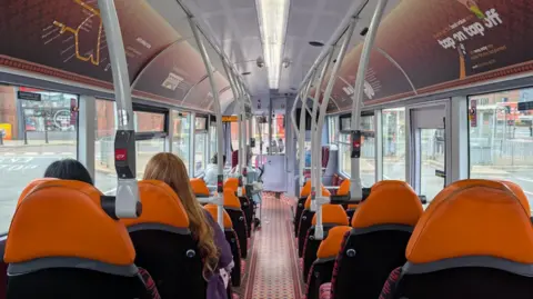 View is from the back of a bus, with the gangway down the middle. The seats either side have orange headrests and there are handle rails down either side. Windows fill each side of the bus and there are two women sat together on one set of seats, one brunette and one blonde and they have their backs to the camera.