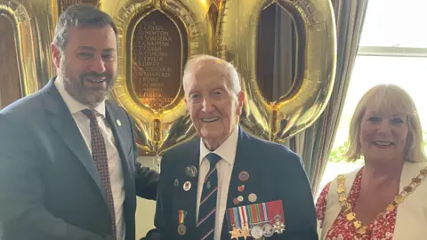 Bath and North East Somerset Council Stan Ford wearing a dark suit, adorned with colourful badges and wartime medals. He is standing beside a man and a woman in a council office, with large gold mylar balloons behind him which read '100'. 