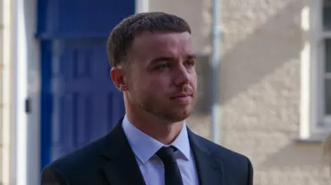 BBC Lewis Peter Carr arriving at Jersey Royal Court for L'Ecume II Trial in 2025. He's wearing a black suit and tie with a cream building and a blue door behind him. 