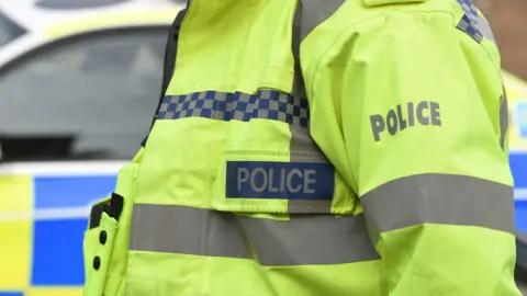 The side profile of a police officers yellow high vis jacket and a police car in the background.