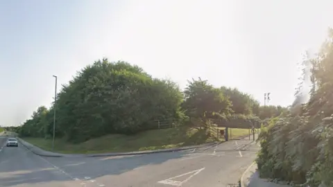 Google Streetview image of a gated entrance to a tree covered area off a road.