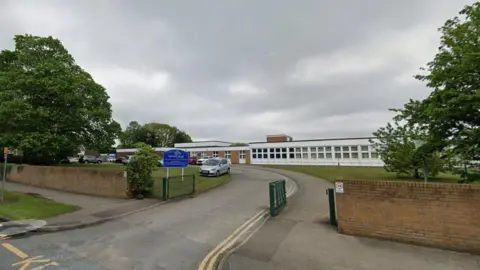 A single-storey building with windows. It is set back from the roadside and there is a gate and wall bordering the site. Outside is a blue sign on the driveway.