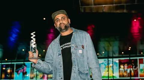 Welsh Music Prize Aly Jamal wearing a black t-shirt and blue denim jacket, and a hat. He is looking at the camera and holding the award which is white and silver. 