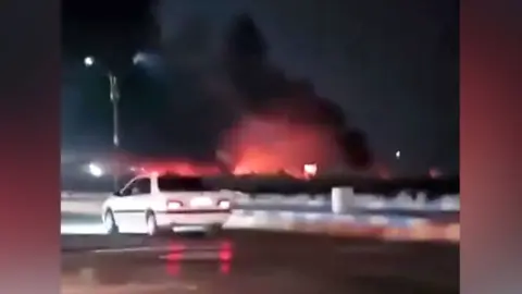 A large fire blazes in the distance beyond a road with a car.