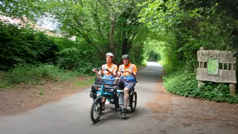 Milestones Trust Edouard and John cycling on a side-by-side tandem along an old railway path between woodland with a sign saying Warmley Forest in the background