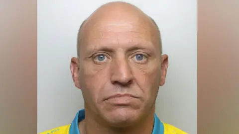 West Yorkshire Police A bald man in his 40s, wearing a yellow top with a blue collar which looks like a football shirt. He is staring into the camera.