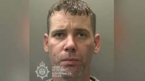 South Wales Police A police mugshot of Christopher West. He has brown hair and stubble and is looking straight at the camera. There is a grey background and the South Wales Police badge is visible.