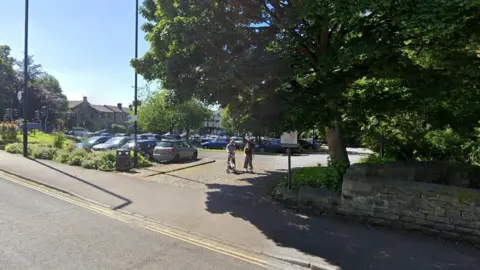 Google A family exits the entrance to a car park in Horsforth Leeds. More cars are in the parking lot as well as more trees.