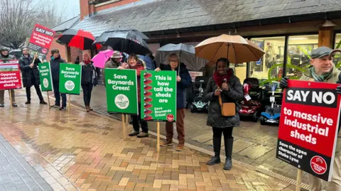 Protesters stand with umbrellas in the rain ahead of the planning meeting. They are holding red and green placards. One says "SAY NO to massive industrial sheds" and another says "Save Baynards Green!".