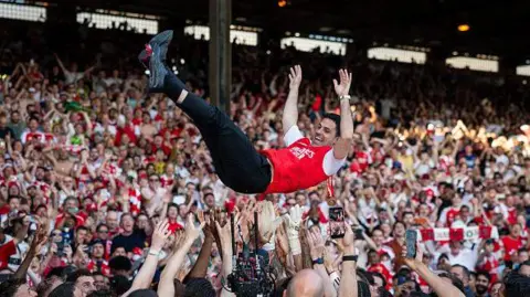 Getty Mikel Arteta is thrown into the air by his players in front of a packed crowd of Arsenal supporters, arms outstretched mid-celebration.