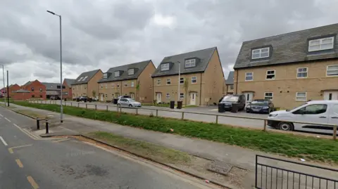Google A row of residential houses on Preston Road in Hull. Cars are parked in front of them. There is a wooden fence, a grass verge, a path and then a road in front of the houses. The sky is overcast. 