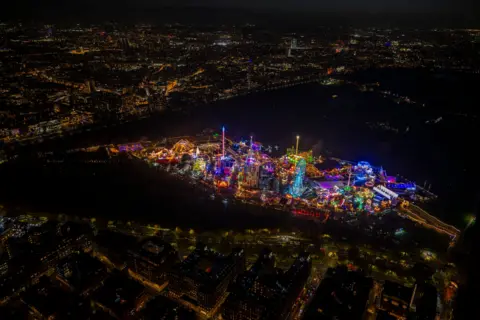 jasonhawkes.com A very large fairground with many attractions such as carousels are lit up in many colours. At the centre of the image is a large ferris wheel lit up in blue. The attraction is pictured from a distance.