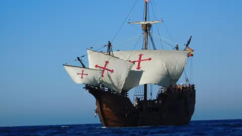 The Nao Victoria Foundation A replica of Christopher Columbus's flagship, The Nao Santa María. It is a tall wooden ship with four masts, five sails, and five decks. The sails are white with a red cross. It is sailing.