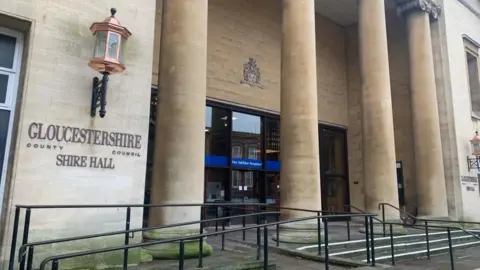 The exterior entrance to Shire Hall in Gloucestershire. It shows a sand coloured building with four large pillars outside, and a large sconce lantern. 