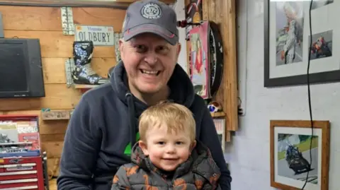 Supplied Trevor is pictured wearing a baseball cap and black hoodie with his son, a young boy with blond hair