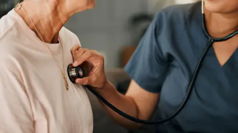 Cardiology consultation, doctor and senior woman consulting about healthcare on living room sofa in retirement home. Hand of nurse with stethoscope while helping elderly patient with medical service - stock photo.