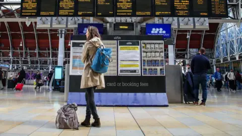 A woman in a brown trench coat with blue and brown bags waits in front of a Great Western Railway ticket machine and arrivals boards on the concourse at Paddington Station. Other travellers with luggage move around in the background