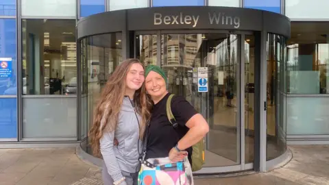 Lizzie Pitt Two women stand outside the Bexley Wing entrance, near a revolving glass door and hospital signage.