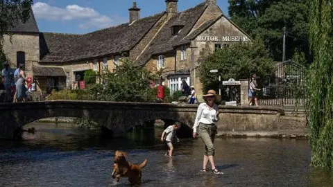 Getty Images A picture of a woman, child and a dog walking in a shallow stream with a bridge and building in the background. The woman is wearing a sunhat and she has the legs of her khaki trousers rolled up. The dog is running happily through the water.