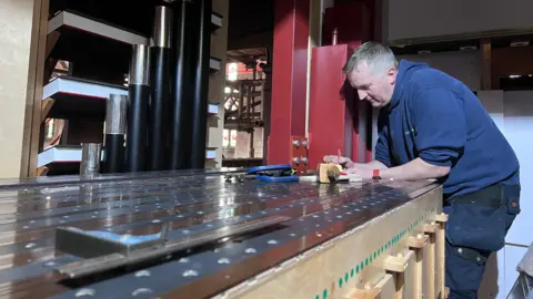 A craftsperson works on part of a large organ mechanism, leaning over a metal surface surrounded by wooden framing and organ pipes.