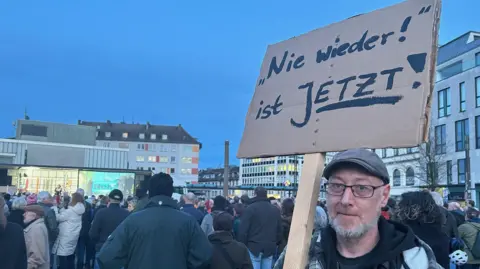 A man with a beard and a flat cap holds up a sign in German that reads "Never again is Now"