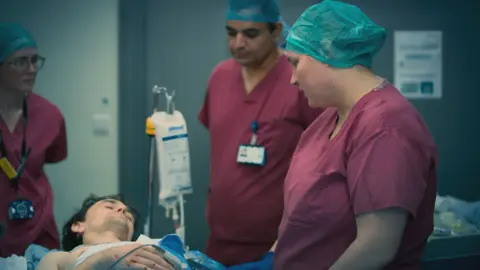 BBC/ExpectationTV Three healthcare staff standing around a male patient on a hospital bed. The patient appears to be on an intravenous drip, with a wire attached to his finger and a strap across his chest.