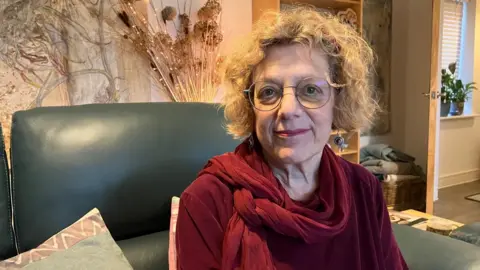 A woman wearing round glasses and red lipstick smiles at the camera. She's also wearing dangly earrings, a velvet red top and scarf. Behind her is an example of her husband's art work on the wall.