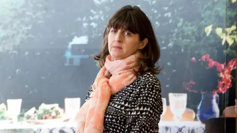 PA Media A woman with dark hair, a pink scarf and a black and white top, stands in front of a canvas of a garden.