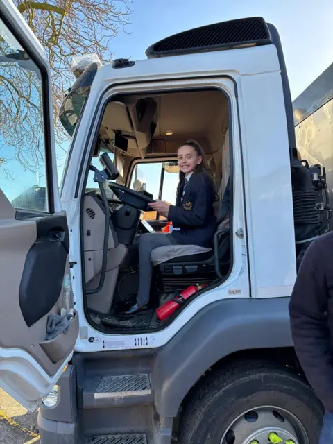 East Lindsey District Council One of the girls, Brooke, sitting inside the cab of a road sweeper. The front door is open. She is holding the steering wheel and smiling at the camera.