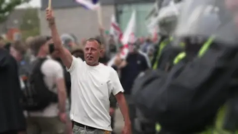 BBC An image of Stephen Roughley in a white t-shirt, holding an item aloft in his right hand at a riot. 