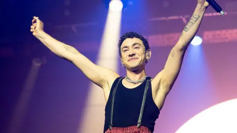 A man on stage. He is smiling with his arms in the air. The lighting has a purple hue. 