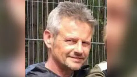 Chris Davies smiles at the camera. He has short grey hair and is wearing a dark coloured top. He is standing in front of some metal fencing.