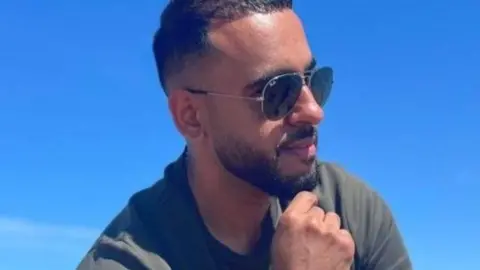 A man with dark hair and a beard is wearing sunglasses. Blue sky is behind him.