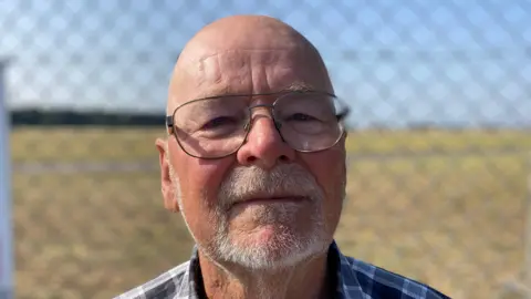 Luke Deal/BBC John Millard smiles at the camera. Behind him, behind a fence, is RAF Lakenheath's base. He is bald and wears glasses with a blue and white chequered shirt.