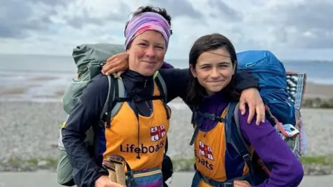 Kerry-Anne Kerry and Finn selfie smiling for the camera as they walk in Wales, each wearing a backpack and the top of a yellow RNLI lifeboat.