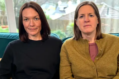 Two women sit indoors on a sofa. A window behind them shows an outdoor scene with trees and rooftops in soft focus.