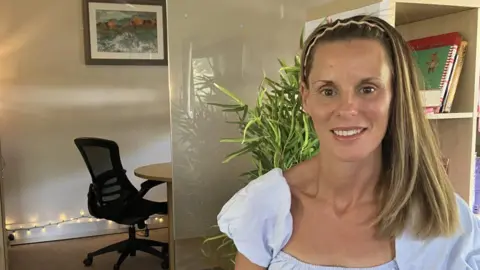 A woman with blonde hair looks at the camera, with a plant and office furniture behind her.