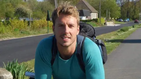 Dan Smith Dan Smith in a blue top and wearing a back pack while sitting on his bike on a paved area beside a road
