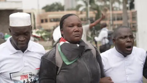 Jacinta Anyango, wearing a black jumper and a Kenyan flag around her neck, stands between two men